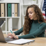 Fiatal nő tanul angolul laptopon keresztül, jegyzetel amerikai zászló előtt