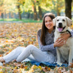 Fiatal nő ölében egy labrador kutyával, őszi falevelekkel borított parkban, mosolyogva pózolnak