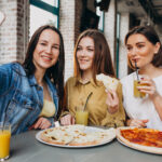 Három fiatal nő pizzát eszik és szelfit készít egy budapesti étteremben, frissítő italokkal és napfényes enteriőrrel