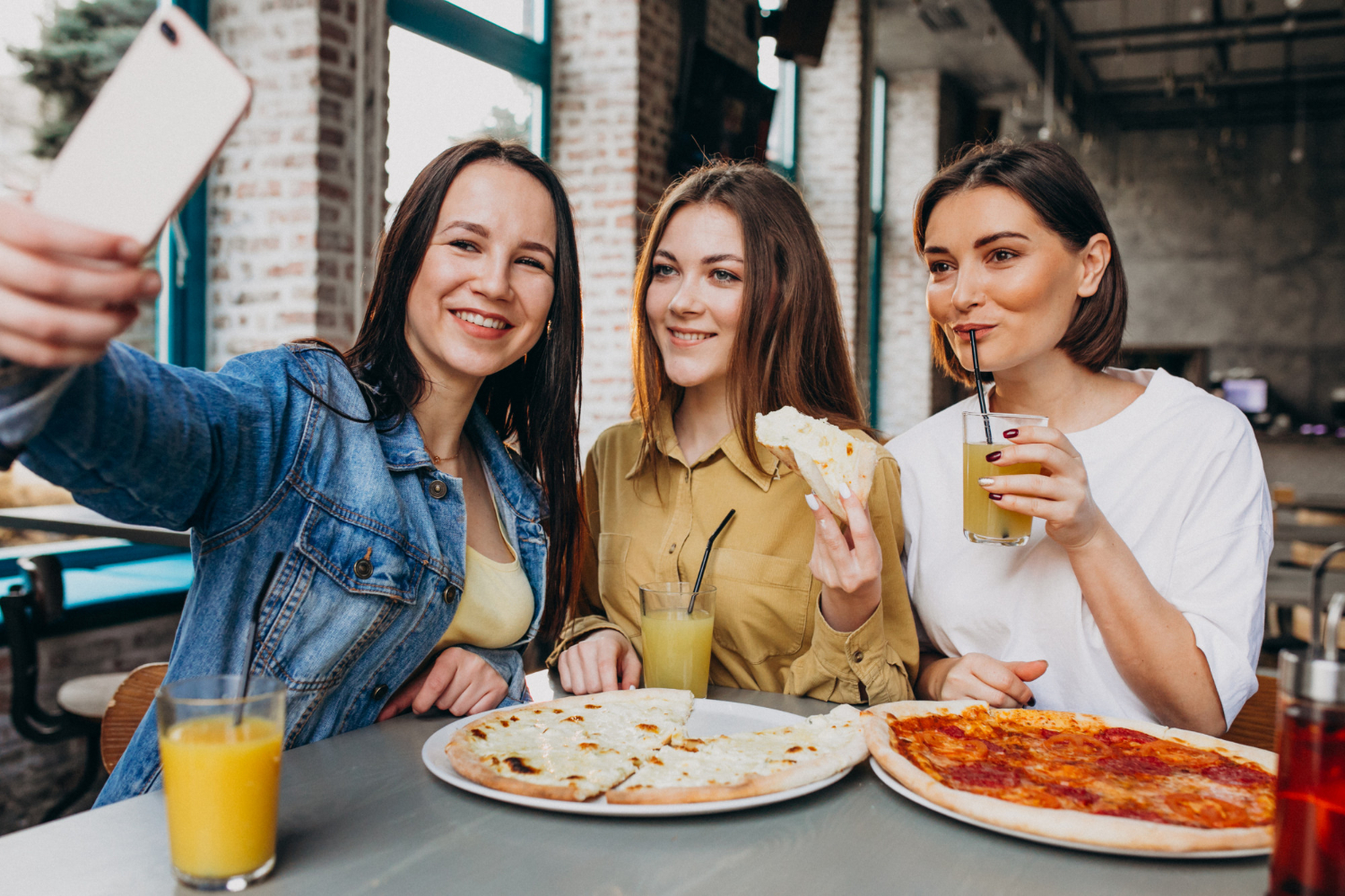 Három fiatal nő pizzát eszik és szelfit készít egy budapesti étteremben, frissítő italokkal és napfényes enteriőrrel